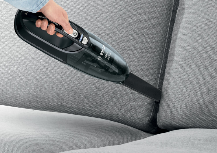 Persona usando una aspiradora de mano Bosch para limpiar las ranuras de un sofá gris