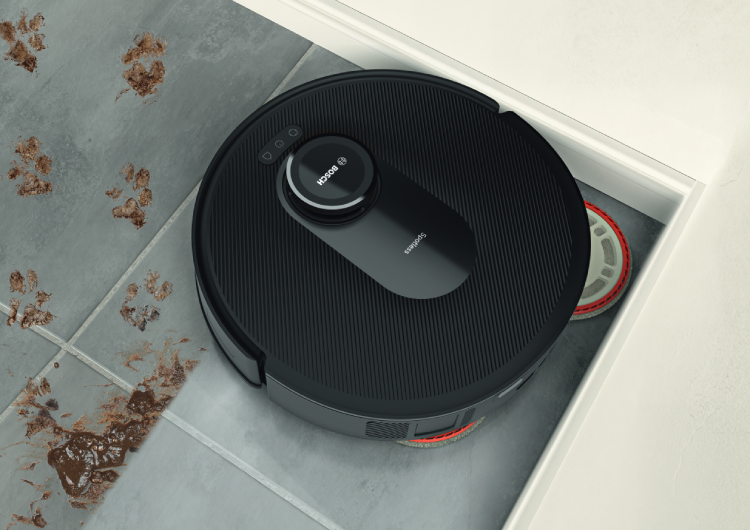 Primer plano de un robot aspirador negro de Bosch limpiando manchas de barro en suelo de baldosa