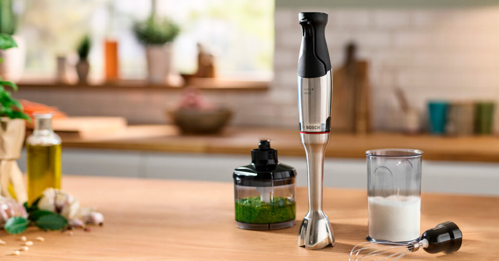 Batidora de mano Bosch sobre encimera de cocina con procesador de alimentos, varilla y vaso mezclador.
