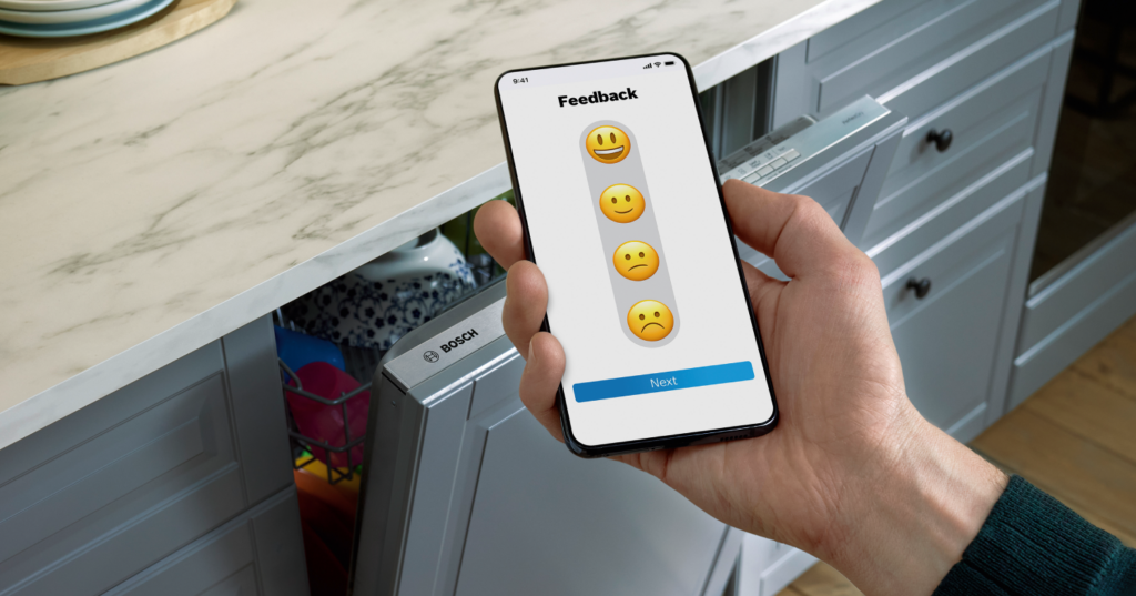 Cocina moderna con lavavajillas Bosch. En primer plano, una mano sosteniendo un smartphone con la aplicación de Home Connect abierta.