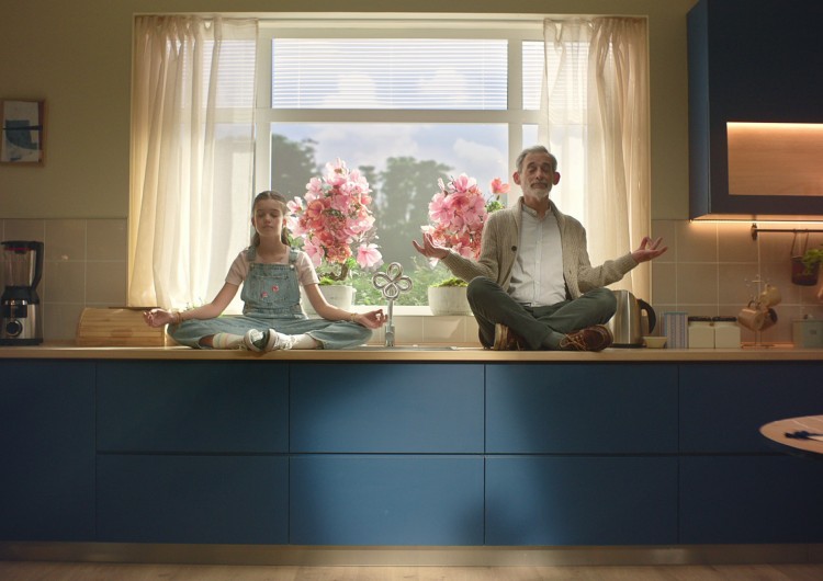 Un abuelo y su nieta sentados meditando sobre la encimera de una cocina azul con un ventanal amplio y flores rosas al fondo