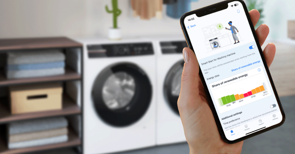 Mano sujeta un móvil en primer plano con la aplicación Bosch Home Connect abierta. Al fondo, lavadora y secadora Bosch.