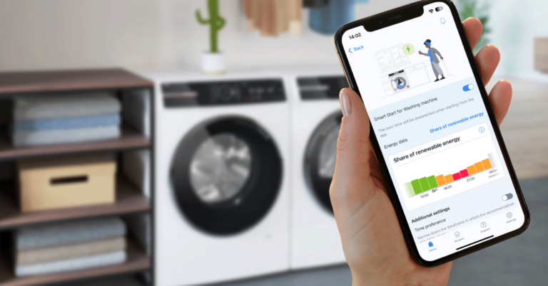 Mano sujeta un móvil en primer plano con la aplicación Bosch Home Connect abierta. Al fondo, lavadora y secadora Bosch.